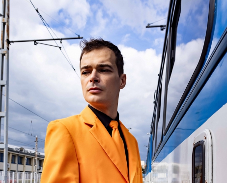 Młody mężczyzna w pomarańczowym garniturze stoi przed wagonem tramwaju i wysuwa palce w celu wciśnięcia guzika otwierającego drzwi. Mężczyzna patrzy w bok. W tle błękitne niebo z białymi, kłębiastymi chmurami.