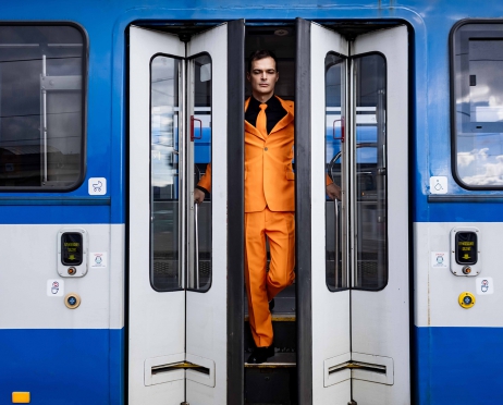 Fragment wagonu tramwajowego w kolorze niebiesko-białym. Na środku drzwi rozsunięte w dwie strony. Pomiędzy drzwiami na schodach młody mężczyzna w pomarańczowym garniturze.