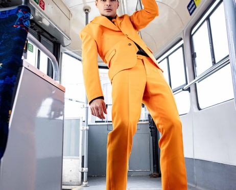 Na pierwszym planie młody mężczyzna w pomarańczowym garniturze i czarnych, lakierowanych butach. Stoi w pustym wagonie tramwajowym, trzyma się za wiszący uchwyt. Wygina się ciałem do przodu, stoi na piętach, patrzy w obiektyw.