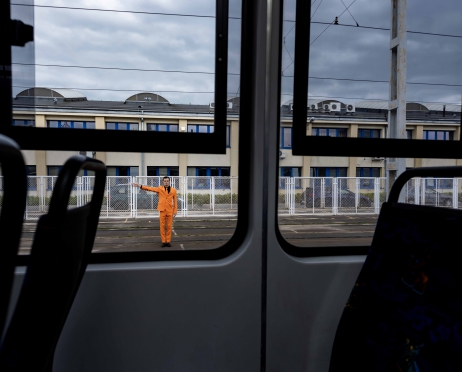 Zdjęcie z wnętrza wagonu tramwajowego. Na pierwszym planie siedzenia i okno tramwaju. Za oknem, na ulicy stoi w dal postać mężczyzny, w pomarańczowym garniturze. Prawą rękę ma wyciągniętą prosto w bok. Patrzy w okno tramwaju. W tle siatka ogrodzeniowa i budynki.