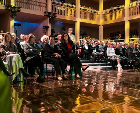 Zdjęcie widowni Teatru Szekspirowskiego w Gdańsku. Ujęcie na salę pełną widzów. Siedzą na krzesłach i na balkonach ze szczebelkowymi bareirkami. Podłoga obita jest złotą podłogą baletową, która daje refleksy dodatkowego światła na widzów i salę.