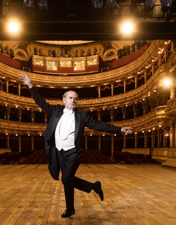 Zdjęcie zrobione ze sceny w kierunku widowni Teatru Słowackiego. Na środku sceny mężczyzna – Jan Peszek, ubrany we frak. Stoi na jednej nodze, drugą nogę ma zgiętą, ręce unosi w górę. Widownia teatru jest rozświetlona i pusta.