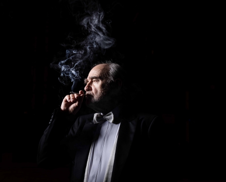 Zdjęcie aktora Jana Peszka. Aktor jest widoczny od pasa w górę, stoi na czarnym tle,  jest ubrany w czarny frak, białą koszulę i biały frak. Wkłada papierosa do ust, nad nim unosi się papierosowy dym. Jasne, białe światło jest tylko na twarzy aktora. Ma krzaczaste brwi.