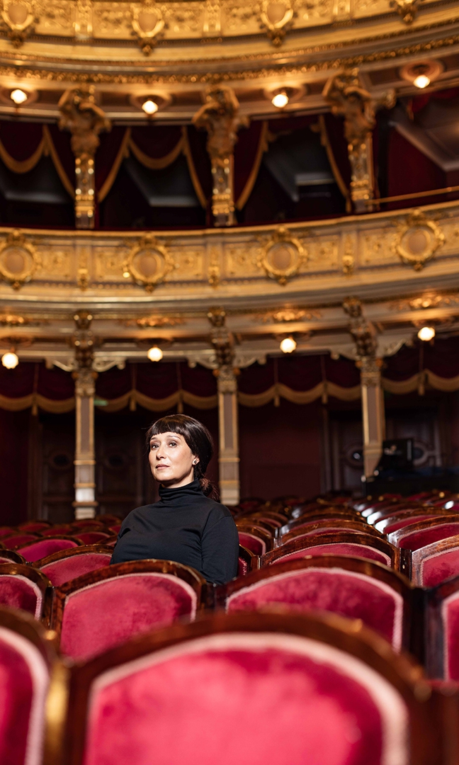 Na zdjęciu brunetka ubrana w czarny golf. Siedzi na fotelu widowni Teatru im. Juliusza Słowackiego w Krakowie. Za nią bogato zdobione balkony lóż widowni.