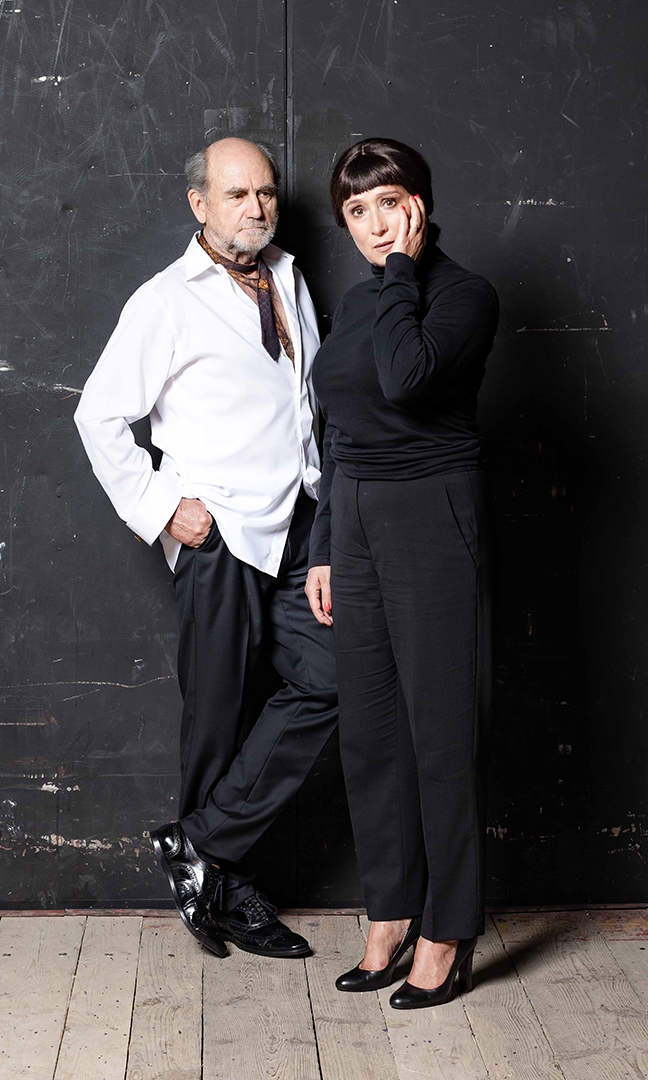 Na zdjęciu na tle czarnej ściany stoi aktor Jan Peszek i aktorka Dominika Bednarczyk. Mężczyzna jest ubrany w luźną, wypuszczoną ze spodni, rozpiętą, białą koszulę. Na szyi ma luźno i niechlujnie zawiązany krawat, czarne spodnie od garnitury i czarne, eleganckie buty. Patrzy na kobietę. Kobieta jest ubrana w czarne spodnie i czarny golf. Ma czarne włosy spięte z tyłu z krótką grzywką do połowy czoła. Ma zatroskaną minę, na policzku trzyma dłoń.