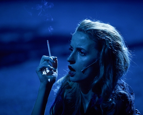 Zdjęcie w niebieskim świetle. Ujęcie obejmuje lewy profil twarzy kobiety, która pali papierosa. Jest rozchochrana i niedbale pomalowana. Patrzy pustym wzrokiem przed siebie. W prawej dłoni na wysokości twarzy trzyma dymiącego, cienkiego papierosa.