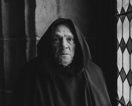 Czarno-biały portret Feliksa Szajnerta w habicie zakonnika. Stary aktor patrzy przestraszony w kierunku obiektywu. Stoi na tle starego muru z witrażem. Na głowie ma kaptur.