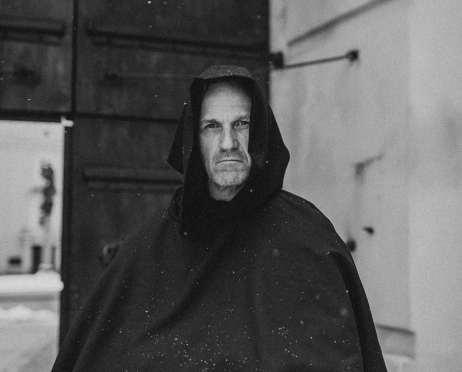 Czarno-biały portret Grzegorza Łukawskiego w habicie zakonnika. Aktor ma na głowie kaptur i dłonie schowane w szerokich rękawach habitu. Stoi na tle otwartych drzwi do dziedzińca zabytkowych zabudowań.