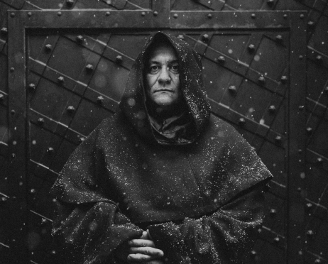 Czarno-biały portret Macieja Jackowskiego w habicie zakonnika.  Aktor z poważną miną stoi na tle zdobionych, metalowych drzwi. Ma na głowie kaptur i dłonie schowane w szerokich rękawach habitu Zdjęcie wykonano na zewnątrz, w  powietrzu unoszą się płatki śniegu.