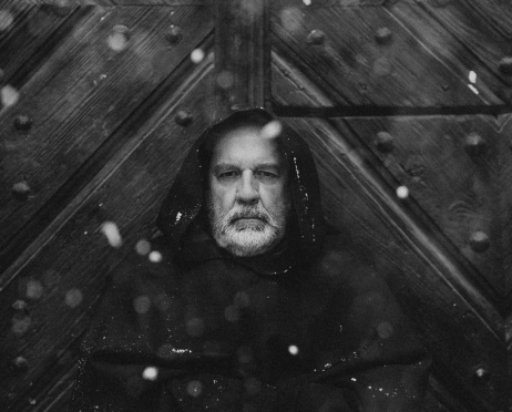 Czarno-biały portret Tomasza Międzika w habicie zakonnika. Stary aktor z poważną miną stoi na tle dużych drewnianych drzwi. Ma na głowie kaptur i dłonie schowane w szerokich rękawach habitu. Zdjęcie wykonano na zewnątrz, w powietrzu unoszą się płatki śniegu.