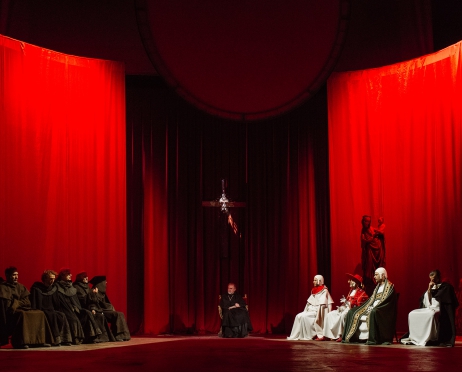 Grupa zakonników i księży siedzi w półokręgu. Scena oświetlona na czerwono. Z lewej strony na ławie siedzi grupa zakonników. W centralnym miejscu w fotelu siedzi ksiądz, za nim wisi krzyż. Po prawej stronie księża w białych sutannach i kolorowych, ozdobionych ornatach.