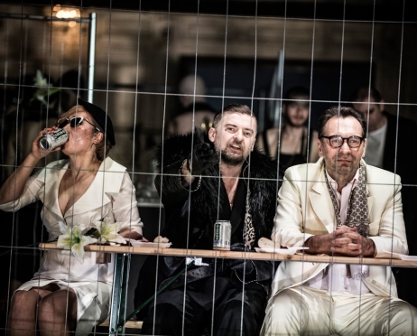 Na zdjęciu trzy osoby siedzą przy piknikowym stole. Pierwsza z lewej siedzi kobieta w białej garsonce i ciemnych okularach, obraca się w lewą stronę pijąc piwo z puszki. Obok niej mężczyzna w czarnym futrze, wskazuje ręką przed siebie. Obok niego mężczyzna w białym garniturze i okrągłych korekcyjnych okularach patrzy zrezygnowany przed siebie. Bohaterowie zdjęcia siedzą za kratami metalowej siatki.