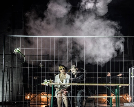 Przy piknikowym stole siedzi kobieta i mężczyzna. Ona jest ubrana w białą garsonkę, kapelusz i ma ciemne okulary. Bezradnie siedzi zwrócona w obiektyw. On w czarnym futrze pochyla się w jej stronę. W tl unosi się dym. Na stole leżą puszki po piwie i biała, sztuczna lilia. Bohaterowie zdjęcia są za metalową siatką ogrodzeniową.
