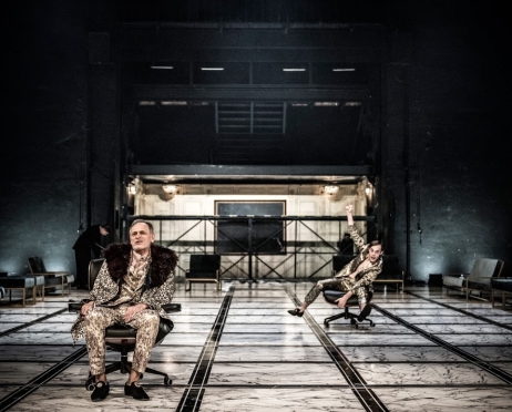Duża Scena Teatru, na podłodze biała, marmurowa podłoga w czarną kratę. Na pierwszym planie dwóch mężczyzn w identycznych garniturach. Z lewej, w fotelu na kółkach siedzi starszy. W głębi na obrotowym podnóżku siedzi młodszy. W lewym rogu pochylony mężczyzna w czarnym mundurze. W głębi widać szyb windy towarowej i zabudowaną na złoto windę.