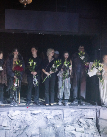 Obsada i twórcy spektaklu „Koriolan” stoją w jednym rzędzie na scenie Domu Machin w Teatrze Słowackiego, podczas ukłonów. Każda z osób trzyma w rękach bukiet, długie, eleganckie kompozycje z białych lilii i zielonych liści. Scenografia ma surowy, postapokaliptyczny charakter: na pierwszym planie widoczna jest zniszczona, pokruszona powierzchnia przypominająca beton lub kamień, z fragmentami gruzu, porozrzucanymi elementami i reliefami sugerującymi ruinę lub upadek miasta. Z boku widać metalowe konstrukcje i techniczne elementy sceny. Tło pozostaje ciemne.