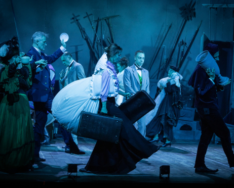 Na dużej scenie Teatru imienia Juliusza Słowackiego w Krakowie około 10 aktorów z obsady spektaklu „Kraków narodowej sztuce, czyli tryumf miernoty” stoją na scenie w intensywnym, niebiesko-turkusowym świetle. Aktorzy mają na sobie kostiumy inspirowane przełomem XIX i XX wieku, zróżnicowane pod względem kroju i zdobień. Na pierwszym planie, pośrodku, kobieta w obszernej, jasnej sukni z falbanami i fioletowymi rękawami pochyla się do przodu, trzymając w obu dłoniach duże, czarne walizki. Po lewej stronie stoi kobieta w długiej, zielonej spódnicy, ciemnym gorsecie i ozdobnym kapeluszu z piórami. Obok niej mężczyzna w granatowym garniturze unosi rękę z białym talerzykiem, a kolejna postać trzyma porcelanowy imbryk. W głębi widać aktorów w jasnych i ciemnych garniturach oraz kostiumach stylizowanych na stroje z epoki. Po prawej stronie drobna postać w bogato zdobionym stroju stoi na schodach, a skrajnie po prawej mężczyzna w bordowym welurowym, ubraniu stańczyka niesie na ramieniu duży, niebieski przedmiot przypominający urnę lub wazę. W tle stoją liczne długie halabardy, włócznie i inne rekwizyty, a drewniana podłoga sceny rozświetlona jest chłodnym, niebiesko-turkusowym światłem.
