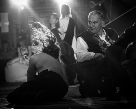 Bartosz Roch Nowicki po lewej stronie, ukazany w czarno-białej tonacji, gwałtownie odchyla tułów do tyłu, znajdując się nisko nad drewnianą podłogą dużej sceny Teatru imienia Juliusza Słowackiego w Krakowie. Jego długie, kręcone włosy unoszą się i rozwiewają wokół głowy, podkreślając dynamikę ruchu. Ma nagie nagie plecy i ramiona, a dolną część ciała okrywa ciemna tkanina. Po prawej stronie Maciej Jackowski, starszy mężczyzna o krótko ostrzyżonych, siwiejących włosach, siedzi na drewnianym krześle. Ma na sobie strój ludowy, jasną koszulę z podwiniętymi rękawami i ciemną kamizelkę ozdobioną dekoracyjnymi elementami. Jedną rękę opiera na podłokietniku, a głowę odwraca w lewo, spoglądając w górę z czujnym wyrazem twarzy. W głębi widać kilka rozmytych postaci. Na mężczyzn pada z góry mocne światło, a pozostałe części otoczenia są w cieniu.