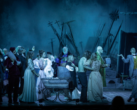 W niebiesko-turkusowym świetle na dużej scenie Teatru imienia Juliusza Słowackiego w Krakowie stoi około 20 aktorów z obsady spektaklu „Kraków narodowej sztuce, czyli tryumf miernoty". Aktorzy mają na sobie kostiumy inspirowane przełomem XIX i XX wiek. W centrum stoi metalowy wózek dziecięcy, z dużymi szprychowymi kołami i podniesioną budką. Po lewej stronie stoi mężczyzna w bordowym, welurowym stroju stańczyka. Mężczyzna niesie na ramieniu duży, szary przedmiot przypominający urnę lub wazę. Obok niego stoją osoby w ciemnych marynarkach, długich sukniach i jasnych bluzkach. Nieco bliżej środka kobieta w długiej, turkusowej sukni z pionowymi pasami trzyma w dłoni niewielki przedmiot, a obok niej znajduje się kobieta w jasnej, liliowej sukni z bufiastymi rękawami i bogatymi falbanami. Za wózkiem stoi siwowłosy mężczyzna w fioletowym stroju kontuszowym, trzymający szablę. Po prawej stronie kilka postaci obejmuje się lub zwraca ku sobie, jedna z nich ma odsłonięty tors i ciemne spodnie, a obok stoi kobieta w długiej, jasnej sukni. W głębi widać łysego mężczyznę w zielonym, ozdobnym płaszczu, trzymającego urnę. Skrajnie po prawej młody mężczyzna w jasnoszarym garniturze, zielonej kamizelce i muszce przyjmuje teatralną pozę z uniesionymi rękami, odchylając głowę i unosząc wzrok ku górze. W tle stoją długie halabardy, włócznie, otwarte trumny i inne rekwizyty.