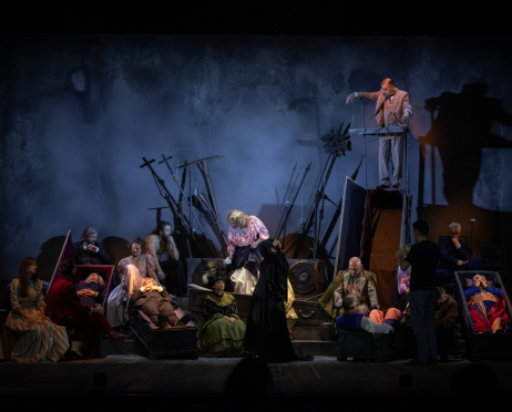 Na dużej scenie Teatru imienia Juliusza Słowackiego w Krakowie, w przygaszonym, niebiesko-szarym świetle znajduje się około 20 osób, ubranych w kostiumy inspirowane przełomem XIX i XX wieku. Po lewej stronie kobieta w jasnej, beżowej sukni siedzi obok młodej kobiety w szarobrązowej sukience. Nieco dalej mężczyzna w czerwonym stroju pochyla się nad otwartą, wyłożoną fioletowym materiałem trumną, w której leży mężczyzna w ciemnym ubraniu. Obok znajduje się kolejna trumna z leżącym aktorem w brązowym stroju, nad nim siedzi kobieta w jasnoróżowej sukni z bufiastymi rękawami. W głębi po lewej stoją i siedzą mężczyźni w ciemnych garniturach, a jeden z nich ma dłuższe włosy i brodę. W centrum kobieta w różowej bluzce z falbanami pochyla się nad rekwizytami, tarczami i pióropuszami. Z przodu siedzi kobieta w zielonej sukni z czarnymi zdobieniami oraz dużym, ciemnym kapeluszem. Po prawej stronie łysy mężczyzna w ozdobnym zielono-złotym płaszczu pochyla się nad niską skrzynią, w której leży aktor w niebiesko-czerwonym stroju. Obok stoi mężczyzna w czarnej koszulce i ciemnych spodniach, to tłumacz polskiego języka migowego, a dalej siedzi siwowłosy mężczyzna w granatowym garniturze. Skrajnie po prawej w kolejnej otwartej trumnie leży aktor w jaskrawym, czerwono-niebieskim kostiumie ze złotymi zdobieniami. W górnej części sceny, na podwyższeniu, stoi mężczyzna w jasnobrązowym garniturze, opierając jedną rękę o metalową konstrukcję i wyciągając drugą w bok. W tle widać liczne długie drągi, włócznie i metalowe rekwizyty, a także drewniane skrzynie, trumny i podesty.