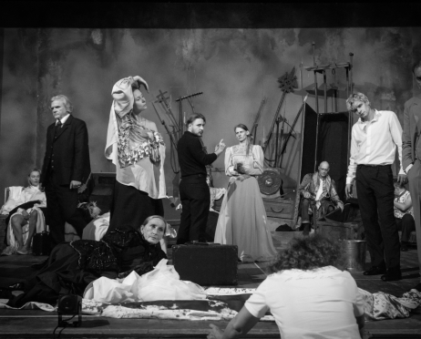 Czarno-biała scena ukazuje obsadę spektaklu „Kraków narodowej sztuce” na dużej scenie Teatru imienia Juliusza Słowackiego w Krakowie. Trzynaście osób znajduje się wśród drewnianych skrzyń, krzeseł i wysokich rekwizytów przypominających broń oraz narzędzia. Aktorzy mają na sobie kostiumy inspirowane przełomem XIX i XX wieku. Po lewej stronie siwowłosy mężczyzna stoi w ciemnym garniturze, a obok kobieta w jasnej, falbaniastej bluzce z nakryciem głowy unosi rękę nad głowę. W centrum mężczyzna w czarnym swetrze, to tłumacz polskiego języka migowego, gestykuluje w stronę kobiety w długiej, jasnej sukni. Po prawej młody mężczyzna w białej koszuli i czarnych spodniach stoi obok starszego mężczyzny w trzyczęściowym garniturze i muszce. W głębi siedzi łysy aktor w ozdobnym stroju. Na pierwszym planie starsza kobieta w czarnej, zdobionej sukni leży na podłodze, a przed nią siedzi tyłem do widza osoba z kręconymi włosami i białą koszulką. Pozostali aktorzy siedzą na skrzyniach lub znajdują się częściowo za innymi postaciami. Tuż pod sceną, na pierwszym planie, stoi reżyserka spektaklu Agata Duda-Gracz, stoi tyłem do nas.