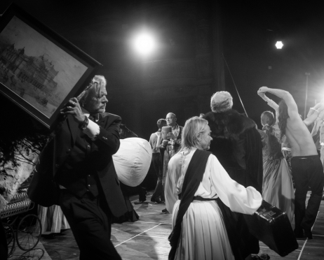 Czarno-biała scena teatralna z udziałem obsady spektaklu "Kraków narodowej sztuce" na dużej scenie Teatru imienia Juliusza Słowackiego w Krakowie. Po lewej stronie Krzysztof Głuchowski, 60 lat, ubrany w ciemny garnitur, trzyma nad sobą duży, ukośnie ustawiony obraz przedstawiający budynek teatru. W głębi, w pobliżu środka, Tomasz Wysocki, 58 lat, stoi wśród pozostałych aktorów i trzyma w dłoniach urnę. Na pierwszym planie, tyłem i częściowo bokiem do widza, Anna Tomaszewska, 75 lat, ma na sobie długą jasną suknię z szerokimi rękawami oraz ciemną szarfę przewieszoną przez ramię. Trzyma czarną walizkę. Pozostali aktorzy noszą między innymi ciemne garnitury, jasne koszule, długie suknie i ozdobne kostiumy. Po prawej stronie mężczyzna bez koszuli unosi ręce nad głową, a obok niego stoi aktor w jasnym garniturze. Mocne światło sceniczne w tle tworzy kontrasty i podkreśla sylwetki postaci na ciemnym tle.