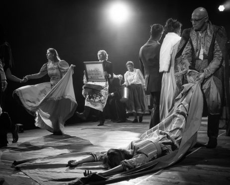 Czarno-biała scena z udziałem obsady spektaklu "Kraków narodowej sztuce" na dużej scenie Teatru imienia Juliusza Słowackiego w Krakowie. Na pierwszym planie Tomasz Wysocki, 58 lat, łysy mężczyzna w okularach ma na sobie bogato zdobiony, wzorzysty kostium z kamizelką i szerokimi klapami. Mężczyzna ciągnie po podłodze Oliwię Durczak, 23 lata, ubraną w jasną suknię w białe pasy. Po lewej stronie Dominika Kozłowska, 29 lat, w długiej sukni z szerokimi rękawami rozkłada materiał i wyciąga rękę w stronę osoby. W głębi Krzysztof Głuchowski, 60 lat, ubrany w ciemny garnitur, niesie oprawiony obraz przedstawiający budynek teatru. Pod obrazem znajduje się wzorzysta, poplamiona tkanina. Za nim Martyna Krzysztofik, 38 lat, ubrana w jasną bluzkę i długą ciemną spódnicę, trzyma metalowe wiadro. W tle widać kolejnych aktorów w eleganckich i teatralnych strojach.