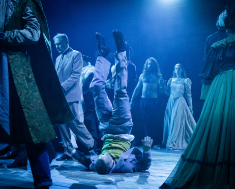 W niebieskim świetle na scenie Teatru imienia Juliusza Słowackiego w Krakowie Wojciech Dolatowski, około 28 lat, leży na plecach na drewnianej podłodze, z nogami uniesionymi pionowo w górę. Ma na sobie jasne spodnie, zieloną koszulę i fioletową marynarkę, ręce rozkłada na boki. Po prawej stronie stoi Dominika Kozłowska, 29 lat, w długiej, jasnej sukni z dopasowaną górą i szeroką spódnicą. Obok niej idzie Bartosz Roch Nowicki, około 40 lat, z nagim torsem i długimi, kręconymi włosami. Po lewej stronie, nieco dalej, znajduje się Rafał Dziwisz, około 60 lat, w jasnym garniturze i koszuli z ciemną muszką, spogląda w dół. Pozostali aktorzy są częściowo widoczni po bokach, w strojach utrzymanych w ciemnych i zielonych odcieniach. Intensywne niebieskie światło oświetla drewnianą podłogę i postacie, tło jest czarne.