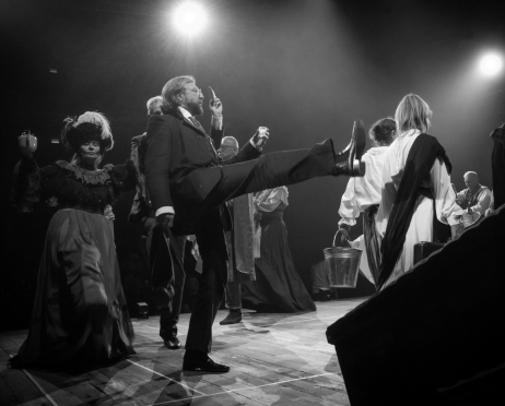 Czarno-biała scena ze spektaklu „Kraków narodowej sztuce” na dużej scenie Teatru imienia Juliusza Słowackiego w Krakowie. Na pierwszym planie, pośrodku, Krzysztof Wrona, 46 lat, w ciemnym garniturze, białej koszuli i krawacie, stoi na jednej nodze i wysoko wyciąga drugą nogę przed siebie. W uniesionej dłoni trzyma mały talerzyk, a głowę odchyla do tyłu. Po lewej stronie stoi Dorota Godzic, 60 lat, w ciemnej, bogato zdobionej sukni z szeroką spódnicą i dużym kapeluszem ozdobionym piórami. W jednej ręce trzyma niewielki imbryk. Dorota Godzic idzie w parze z niewiele widocznym Grzegorzem Łukawskim, 56 lat. Wokół nich znajdują się inni aktorzy w kostiumach, częściowo zasłonięci przez osoby na pierwszym planie. Drewniana podłoga jest mocno oświetlona.