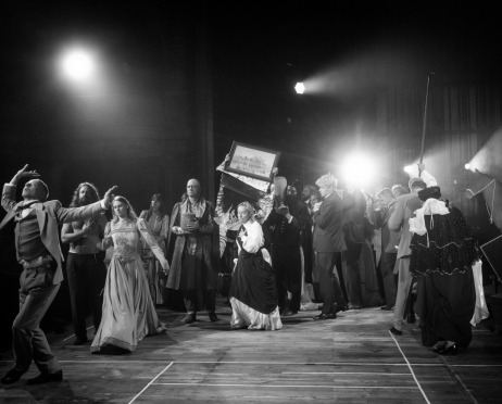 Czarno-biała scena ze spektaklu „Kraków narodowej sztuce” na dużej scenie Teatru imienia Juliusza Słowackiego w Krakowie. Na pierwszym planie, po lewej stronie, Wojciech Dolatowski, 28 lat, w jasnym garniturze, kamizelce i ciemnej muszce, wykonuje dynamiczny gest: odchyla głowę do tyłu, unosi jedną rękę nad głowę, a drugą wyciąga w bok. Za nim stoi Bartosz Roch Nowicki, 37 lat, długowłosy, brodaty mężczyzna z nagim torsem. Aktor idzie w parze z Dominiką Kozłowską, 29 lat, która ma na sobie długą, jasną suknię z ozdobną górą i bufiastymi rękawami. Pośrodku stoi łysy Tomasz Wysocki, 70 lat, mężczyzna w okularach, ubrany w długi, wzorzysty płaszcz; trzyma przed sobą niewielka urnę. Za nim aktorzy unoszą oprawiony obraz przedstawiający budynek Teatru imienia Juliusza Słowackiego, pod którym znajduje się wzorzysta, poplamiona tkanina. Przed obrazem stoi Anna Tomaszewska, 75 lat, w białej tunice przepasanej ciemnym materiałem, z dłonią przyłożoną do piersi. Po prawej stronie zgromadziła się grupa aktorów w zróżnicowanych kostiumach. Na pierwszym planie widać postać odwróconą tyłem, ubraną w ciemny, bogato zdobiony strój z koronkowymi elementami. Drewniana podłoga jest oświetlona reflektorami, z których jeden mocno rozświetla prawą część sceny.