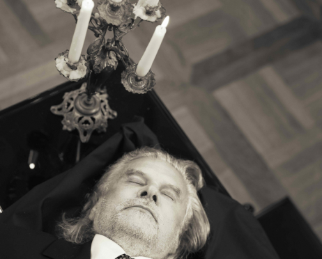 W ciepłych, przygaszonych odcieniach sepii leży na fortepianie Krzysztof Głuchowski, widoczny od głowy do klatki piersiowej. Ma średniej długości, siwe włosy zaczesane do tyłu oraz lekki zarost. Leży na plecach na czarnym materiale, z zamkniętymi oczami i głową odchyloną do tyłu. Ubrany jest w ciemny garnitur, białą koszulę i ciemny, ozdobny krawat.Nad jego głową, w lewej górnej części, znajduje się ozdobny, metalowy świecznik z pięcioma białymi świecami różnej wysokości. Na końcach świec są płomienie. Świecznik ma dekoracyjne ramiona i bogato zdobioną podstawę. Stoi na błyszczącej, czarnej powierzchni fortepianu. W tle widać drewnianą podłogę ułożoną w geometryczny wzór.