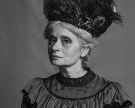 Portret Doroty Godzic z bliskiej perspektywy utrzymany w czarno-białej kolorystyce. Kobieta ma 60 lat, ma na sobie ciemną suknię z szeroką falbaną wokół dekoltu i ramion. Jej siwe włosy są upięte pod wysokim, czarnym kapeluszem ozdobionym licznymi piórami i dekoracyjną broszką. Patrzy lekko w lewo, z poważnym wyrazem twarzy. W tle znajduje się gładka, szara ściana.