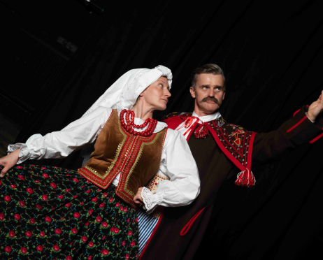 Martyna Krzysztofik, 38 lat, i Marcin Sianko, 51 lat, w tradycyjnych strojach ludowych na tle czarnej kurtyny. Martyna Krzysztofik stoi po lewej stronie. Ma na sobie białą bluzkę z szerokimi rękawami i koronkowymi mankietami, brązowy, bogato zdobiony haftami gorset oraz długą, ciemną spódnicę w czerwone i różowe kwiaty. Na głowie nosi białą chustę, a na szyi kilka sznurów czerwonych korali. Lewą ręką przytrzymuje rozłożoną spódnicę, a głowę odwraca w prawo. Po prawej stronie stoi Marcin Sianko, ubrany w ciemny strój ozdobiony czerwonymi taśmami, kolorowymi haftami i dekoracyjnymi frędzlami. Pod szyją ma białą koszulę z czerwoną wstążką. Unosi prawą rękę w bok. Patrzy w prawo z poważnym wyrazem twarzy. Oboje są lekko odchyleni w przeciwne strony.
