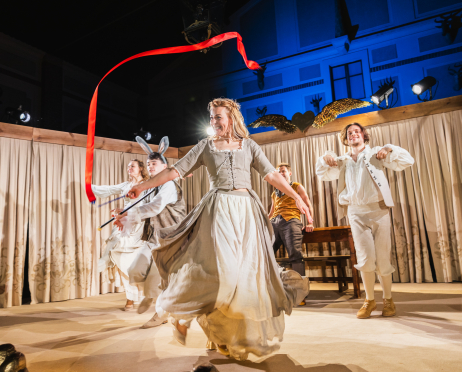 Obsada spektaklu „Kubuś Fatalista i jego Pan” na scenie podczas sceny zbiorowej. Na pierwszym planie aktorka w jasnym, XVIII-wiecznym stroju z rozłożonymi rękami wykonuje sceniczny gest. Wokół niej znajdują się pozostali aktorzy w kostiumach  z epoki.  Po prawej stronie stoi aktor w białym stroju z kamizelką i krótkimi spodniami, a po lewej aktor w białym kostiumie z charakterystycznymi uszami królika, trzymając kij. W głębi sceny widoczny jest Mateusz Marczydło w brązowym stroju. Nad aktorami unosi się długa, czerwona wstęga, podkreślająca ruch i dynamikę przedstawienia. W tle widać udrapowane zasłony, drewniane elementy scenografii oraz reflektory.