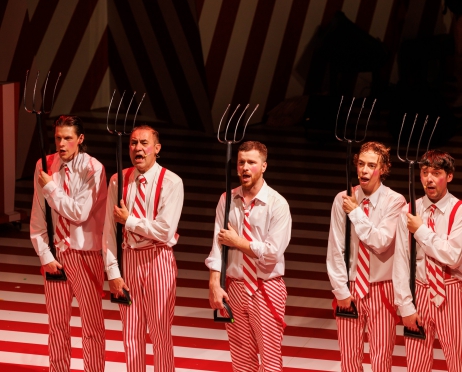 Na zdjęciu w centralnej części na środku pięciu mężczyzn. Stoją obok siebie w rzędzie. Coś mówią wyglądają jakby śpiewali. W ręku każdy z nich trzyma widły wzniesione w górę. Ubrani są w białe koszule, biało czerwone krawaty w paski biało czerwone spodnie w paski. Tło jest czerwono białe.