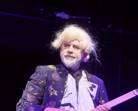 Starszy mężczyzna z peruką na twarzy w stylu dworskim z czasów carskich. Ubrany w fioletowy surdut we zwory i białą koszulę. Patrzy zniesmaczony. W rękach trzyma gitarę.