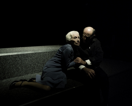 Na zdjęciu Anna Polony i Jan Peszek. Aktorka siedzi bokiem i twarzą wtula się w ramię mężczyzny. On z czułością obejmuje jej dłoń i patrzy na nią Ona jest ubrana w niebieską sukienkę, on w czarny, luźny płaszcz.