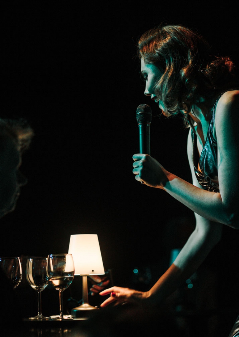 Na zdjęciu aktorka trzymając mikrofon w ręce śpiewa. Ujęcie od boku. Z uśmiechem pochyla się nad stolikiem ze świecąca lampką. Przy stoliku siedzi mężczyzna z kobietą. Aktorka jest ubrana w wieczorową suknię i brązowe włosy uczesane a’la Marylin Monroe. Na zdjęciu panuje półmrok.
