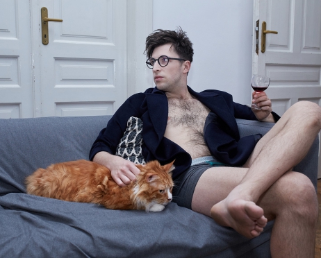 Na zdjęciu młody mężczyzna siedzi na błękitnej kanapie. Jest w bokserkach i marynarce. Prawą ręką głaszcze rudego, puchatego kota, w lewej trzyma kieliszek z winem. Mężczyzna skręca głowę w swoją prawą stronę. W tle białe drzwi.