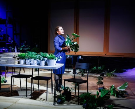 Na zdjęciu kobieta w niebieskiej sukience. W dłoni trzyma zielony kwiat. Wokół niej, na stole i podłodze, ułożone są zielone kwiaty. Kobieta patrzy przed siebie. Tło jest ciemne, mroczne.