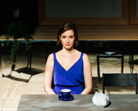 Na zdjęciu kobieta w niebieskiej bluzce siedzi przy stole. Jest spokojna.  Przed nią stoi niebieska filiżanka i biały imbryk. Za nią na stole jest zielona roślina. Tło jest ciemne. Światło pada wyłącznie na dziewczynę.