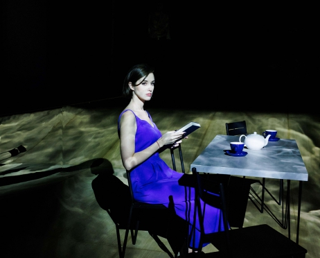 Na zdjęciu dziewczyna w niebieskim spodnium siedzi przy stole. W dłoni trzyma książkę. Spogląda przed siebie. Zdjęcie jest mroczne. Światło pada wyłącznie na dziewczynę i stół.