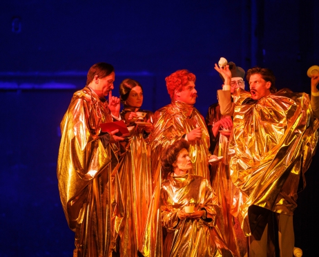 Zdjęcie zbiorowe. Na scenie grupa kobiet i mężczyzn, są ubrani w złote szaty do ziemi, w dłoniach trzymają filiżanki. Część stoi, jedna osoba gestykuluje unosząc wysoko ręce, jedna z osób siedzi. W tle niebieskie światło.