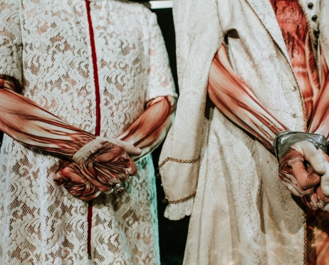 Dwie postaci stoją z rękami złożonymi na wysokości brzucha. Ujęcie ukazuje klatkę piersiową i fragment nóg. Postaci są ubrane w kombinezony z namalowaną strukturą mięśniową  Postać z lewej jest ubrana w białą koronkową tunikę, a druga ma biały kontusz, jej ręce są związane srebrną taśmą klejącą.