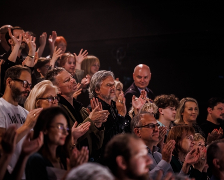 Zdjęcie przedstawia widownię wypełnioną widzami. Grupa osób stoi w rzędach. Klaszczą. Patrzą w kierunku sceny prawdopodobnie na aktorów. Niektórzy są skupieni, inni uśmiechnięci. Są zafascynowani.