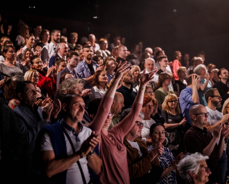 Zdjęcie przedstawia widownię wypełnioną widzami. Grupa osób stoi w rzędach. Klaszczą. Patrzą w kierunku sceny prawdopodobnie na aktorów. Niektórzy są skupieni, inni uśmiechnięci. Są zafascynowani. Tło jest czarne.