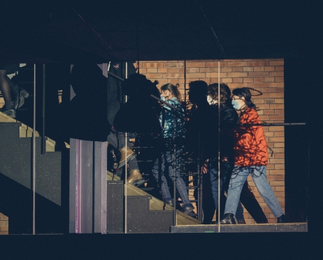 Na zdjęciu grupa osób przemieszcza się po czarnych schodach. Niektórzy zasłaniają twarz maseczkami. Na ich sylwetkach wyświetlana jest projekcja przypominająca kłębek splątanych sznurków. W tle ściana z czerwonej cegły.