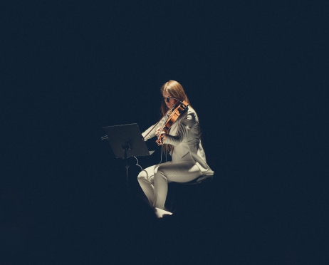 Na zdjęciu dziewczyna, która gra na skrzypcach. Ubrana jest w biały garnitur. Przed nią stoi składany pulpit, na którym są nuty. Wokół mrok. Światło oświetla tylko jej sylwetkę.