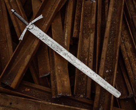 Na dużych, zardzewiałych elementach konstrukcyjnych rozłożonych w nieładzie leży miecz. Miecz jest duży i lekko zniszczony, wygląda jak miecze średniowiecznych rycerzy.