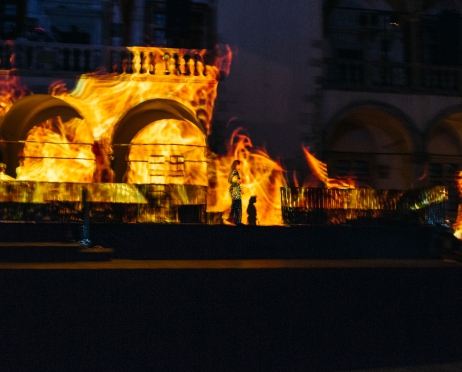 Na murze dziedzińca arkadowego Zamku na Wawelu wyświetlono płomienie. Przez podłużny podest wybudowany wzdłuż muru przechodzi kobieta w cekinowej sukni.