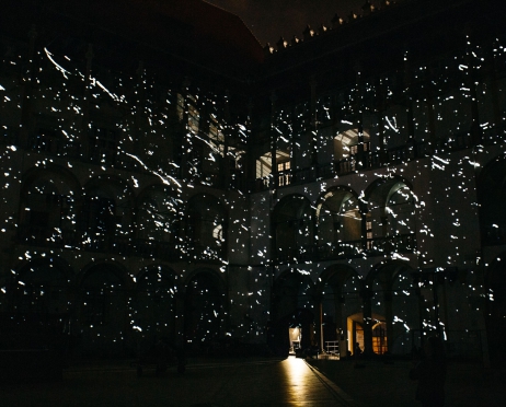 Róg dziedzińca arkadowego Zamku na Wawelu w nocy. Mury oświetlone są projekcją przypominającą rozgwieżdżone niebo. Na dziedzińcu stoją różne elementy scenografii. Z kilku okien bije jasne światło.